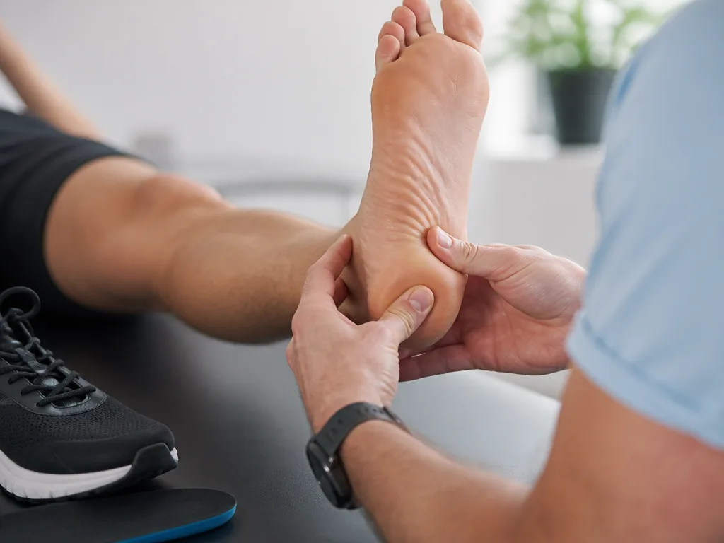 Fysioterapeut undersöker en persons häl och fotsula i en ljus klinikmiljö, med en sportsko och skoinlägg synliga bredvid